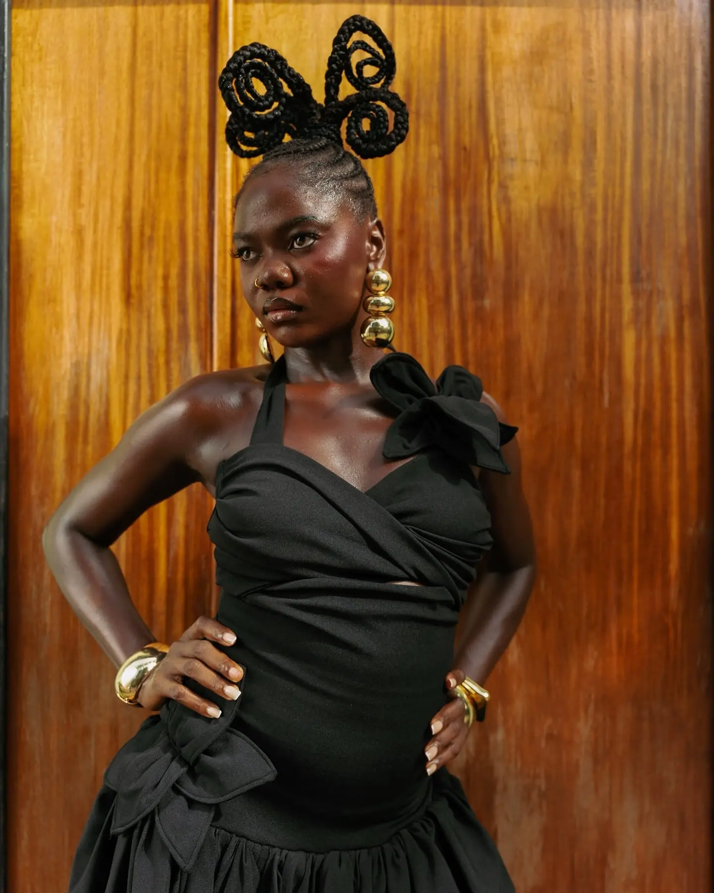 Woman in a black bow-trimmed dress with sculptural looped braids and gold earrings poses hands-on-hips before polished wood doors.