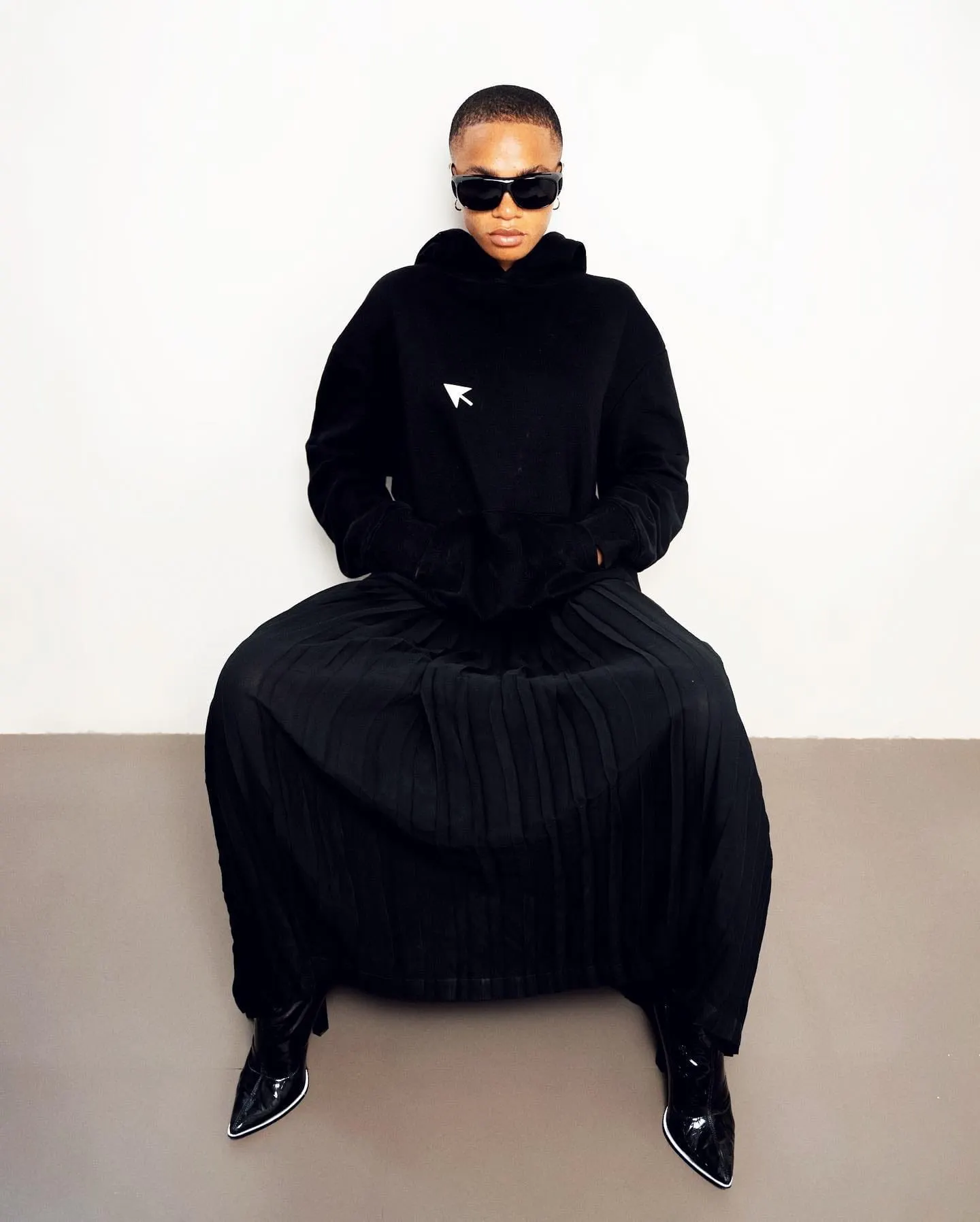 Seated figure in dark sunglasses, oversized black hoodie and wide pleated skirt, pointed boots peeking out.