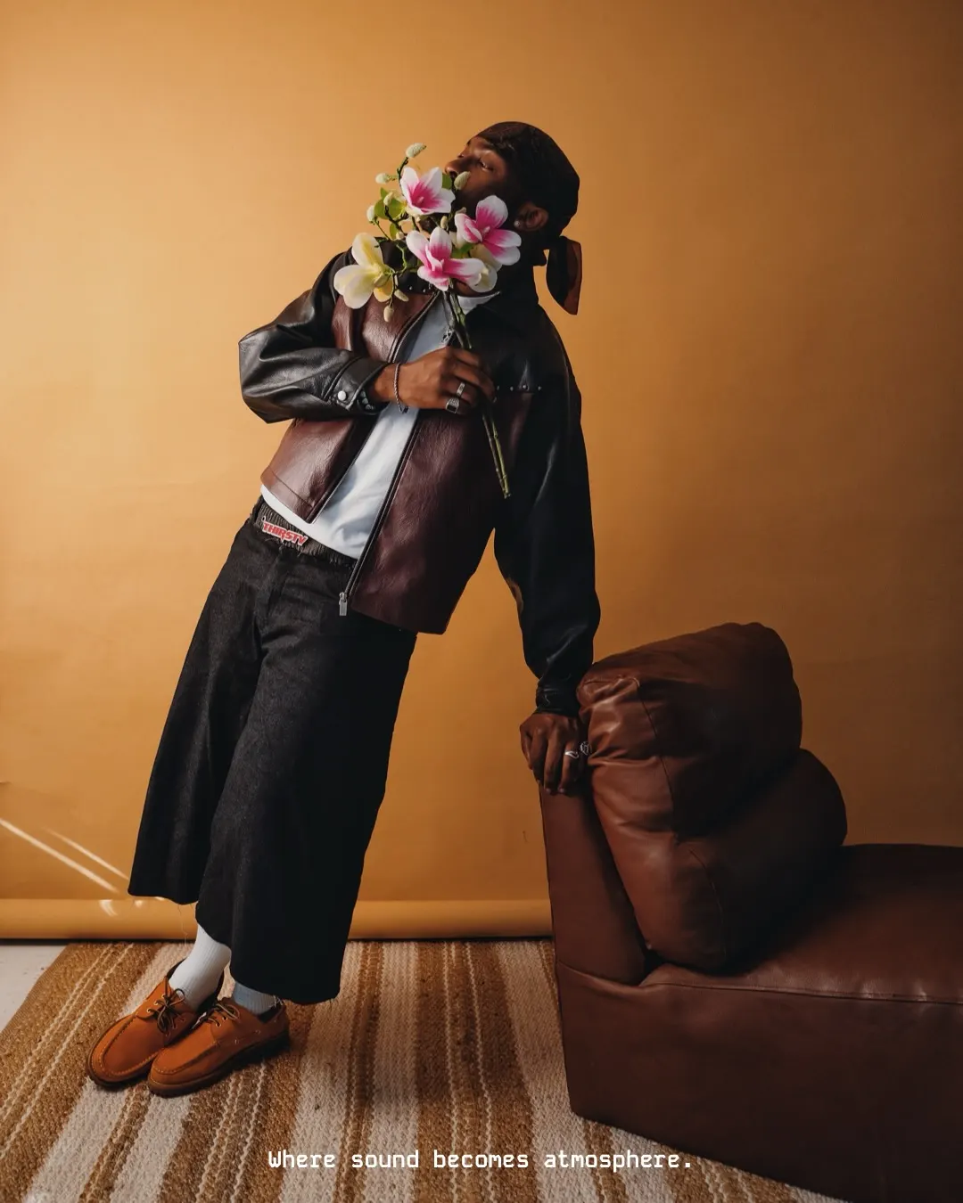 Musician in an oxblood leather jacket and wide dark denim leans back against a brown leather ottoman, head tilted into a bunch of magnolias, on an amber set.