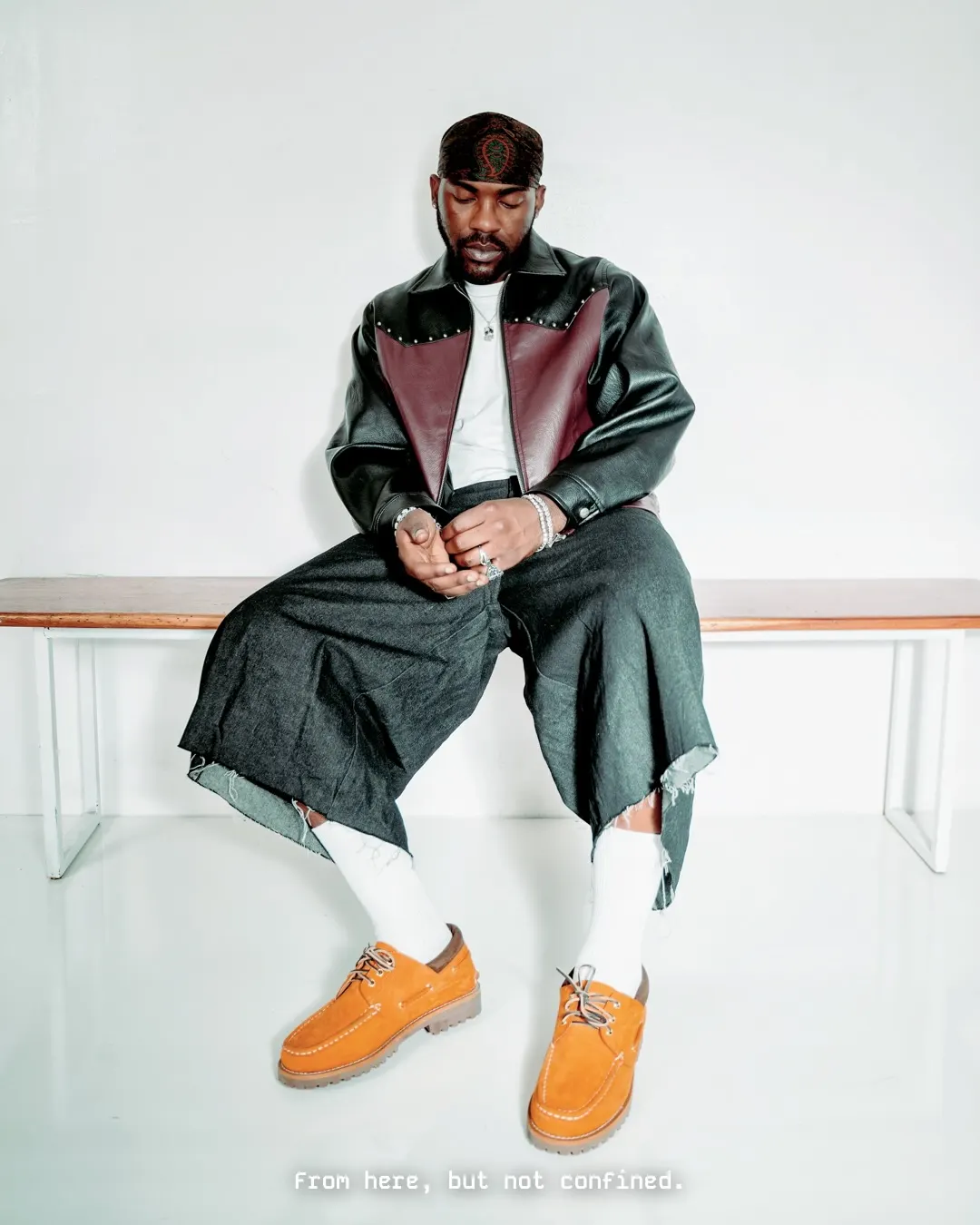Musician in a two-tone leather jacket, frayed wide-leg denim and tan suede shoes sits slumped forward on a low wooden bench in a white studio.