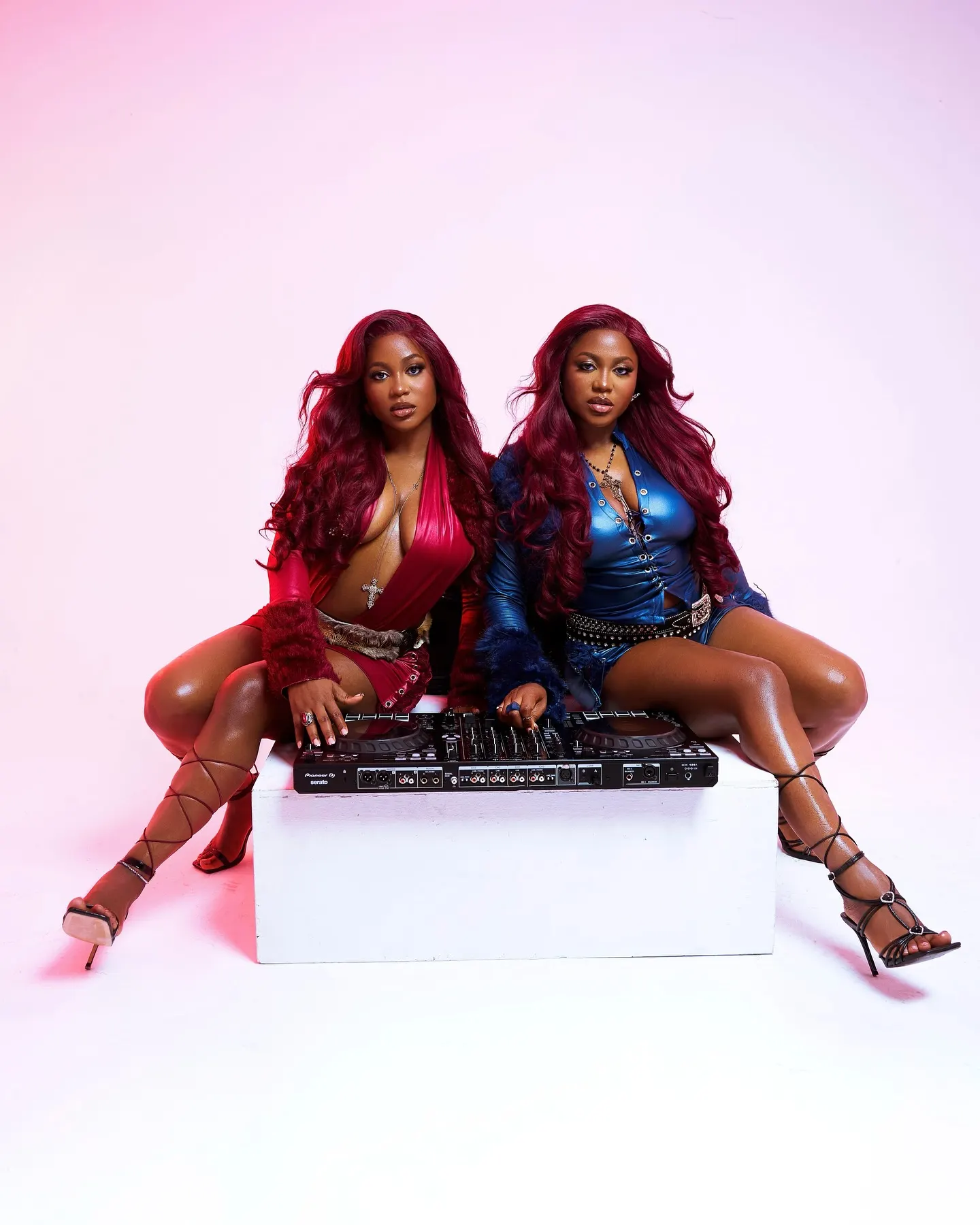 Twins in red and blue leather sit atop a white plinth behind a DJ controller, legs laced into strappy heels against a pink haze.